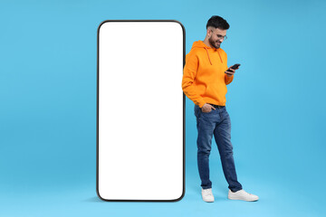 Man with mobile phone standing near huge device with empty screen on light blue background. Mockup...