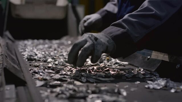 Worker sorts trash on conveyor belt at waste recycling plant. Metal pieces falling into a box from recycling processing machine. Man sorts pieces of metal, iron or steel with his hands