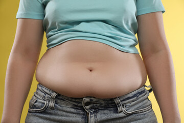 Woman with excessive belly fat on yellow background, closeup. Overweight problem