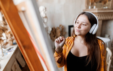 Girl artist paints a picture in a beautiful studio. Female with headphones listening to music.