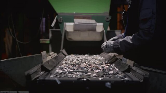 Worker sorts trash on conveyor belt at waste recycling plant. Metal pieces falling into a box from recycling processing machine. Man sorts pieces of metal, iron or steel with his hands