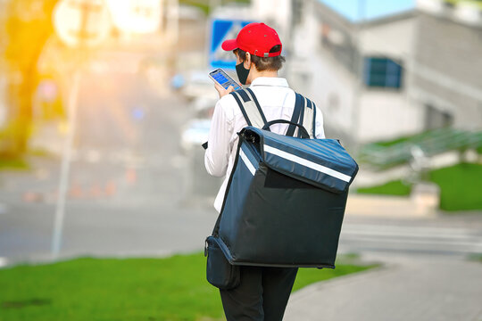 Food delivery boy with black thermal backpack using phone. Boy delivering food orders, food deliver man walking on city street. Deliveryman on the move. Foode delivery service