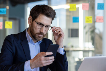 A professional businessman in a suit attentively uses a smartphone, adjusting his glasses in a contemporary office setting with colorful sticky notes in the background. - Powered by Adobe