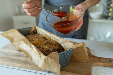 Woman with apron ready to glaze a half cooked meatloaf in a loaf pan. Holding a bowl with ketchup...