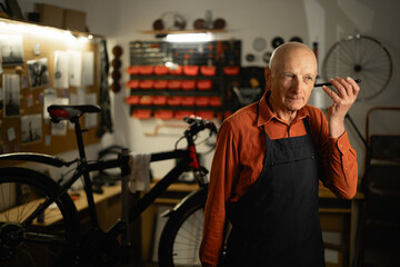 Portrait of old repair man in bicycle shop listening audio message working in cycling workshop and...