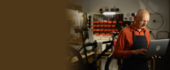 Portrait of senior mechanic worker in his workshop using laptop computer repairing bike. Small business.