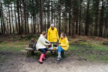 Halt for snack during hiking. Company hikers enjoying picnic on the bench, drinking hot tea, eating sandwiches in forest. Friends relaxing and having snack picnic on nature background. Backpacker trip