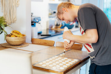 Concentrated young man placing batter on silicone baking sheet to make French macaroons dessert in light modern kitchen at home. Preparing steps for baking cookies. Culinary hobby. Selective focus