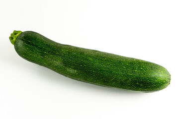 A green zucchini lies on a white background. It has a smooth texture and a slight shine, with a tapered shape and a small stem. The simple setup highlights the vegetable's fresh appearance.