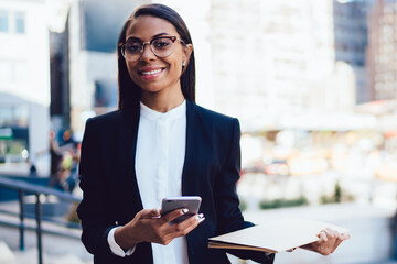 Half length portrait of cheerful african american student of faculty of lawyer dressed in formal wear holding folder and smartphone device in hands standing in downtown in urban setting