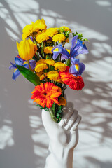 Ceramic Vase made in the shape of human hand with multicolor various summer flower bouquet on white table under sunlight and shadows on gray wall. Trendy interior design decor details. Vertical card.