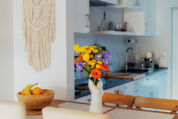 Kitchen counter table with focus on vase made in shape of human hand with multicolor various summer flower bouquet with blurred background of modern cozy white kitchen. Home interior design details.