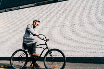 Stylish male in coat, sunglasses and protective helmet riding on retro bicycle near white brick urban wall. Neutral carbon footprint transportation. Green eco friendly mobility sustainable transport.