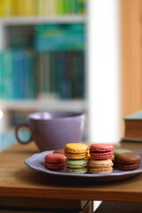 Purple plate filled with pastel macarons, cup of tea or coffee, vintage books and reading glasses on the table. Colorful bookcase in the background. Selective focus.