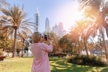 Young tourist woman shooting on mobile phone modern skyscraper in park of Dubai, United Arab...