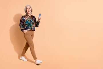 Full length photo of good mood senior person wear print blouse walk to empty space with smartphone...