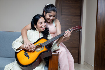 Indian Young Teenage Daughter Helping mother with Guitar Playing at home sitting on sofa, Mother...