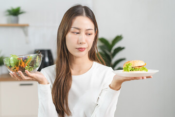 Diet, Dieting asian young woman, girl confused choose, choice eating food, hand holding bowl of vegetable salad and hamburger when hungry in kitchen at home. Temptation of female weight loss person.