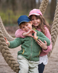 Portrait of a wonderful brother and sister. The girl hugged the boy and folded her hands into a heart. Love in the family. Children play on rope swings outside.