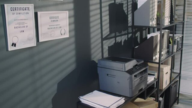 No people tilt footage of multiple certificates of bachelor of law on grey wall and shelf with folders near table with printer in solicitors office in modern law firm