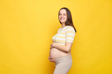 Profile of smiling delighted Caucasian pregnant woman with bare belly wearing casual top isolated...