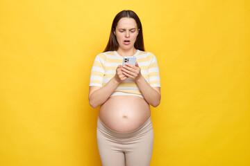 Shocked attractive Caucasian pregnant woman with bare belly wearing casual top holding smartphone...