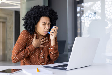 A young woman in a polka-dotted shirt feeling distressed as she uses an inhaler to alleviate asthma symptoms ,while working at her laptop in a sunny office. - Powered by Adobe
