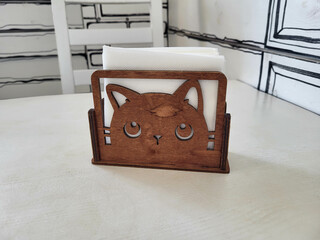 White paper napkins in a brown wooden napkin holder in the shape of a cat's head on a white table.