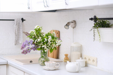 Spring morning in light kitchen: bouquet of white flowers, white teapot and glass of green tea.