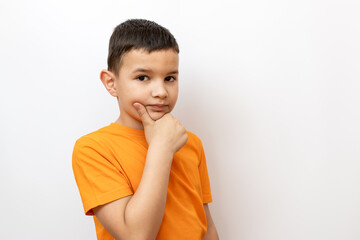 A boy in an orange T-shirt on a white background holds his hand on his chin.