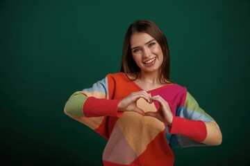 Heart shaped gesture by hands, sending love. Young woman is standing against green background in the studio