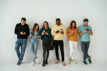 With smartphones in hands. Group of young people are standing against white background