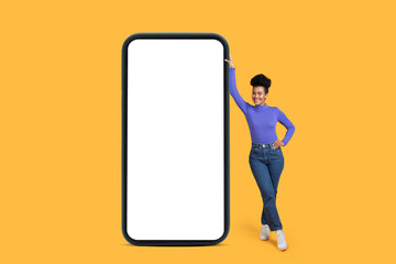 Hispanic woman stands next to a large smartphone with white blank screen isolated on yellow background, mockup copy space