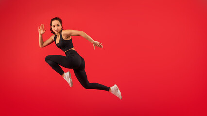 A woman wearing a black top and black leggings is captured mid-jump in the air. Her arms are outstretched and her legs are bent, showcasing her athleticism and agility as she defies gravity.