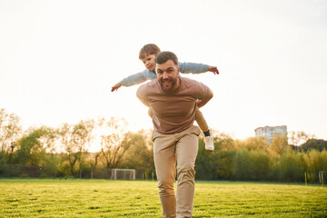 Wellbeing, kid is on the shoulders or back. Happy father with son are having fun on the field at...
