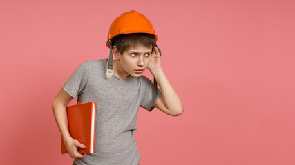 A boy in a construction helmet, a gray T-shirt and a notebook in his hand, listens to something or...