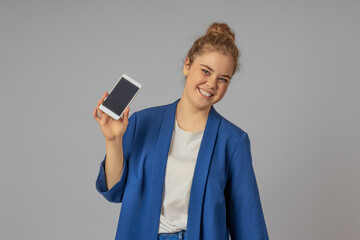 Smiling girl in a blue jacket on a gray background demonstrates the screen of her cell phone