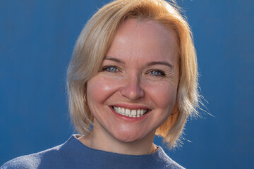 Adult and happy blonde close-up smiles broadly on a blue background in a blue sweater