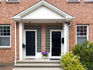 Two black metal doors with flower pots on the steps of duplex houses. It has a red brick wall with white wooden trim. There are glass blocks for windows on both sides of the building entrances.