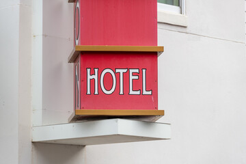 A square hotel sign in the shape of blocks. The signpost rotates on a swivel mechanism on a wall. Each block has a letter for the word hotel. The wooden sign is red with white letters and gold trim.  