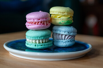 Four French macarons; pink, green, blue, and yellow. The assorted elegant sandwich style sweet dessert is stacked on a blue plate and wooden table. The traditional French pastry has a hard shell.