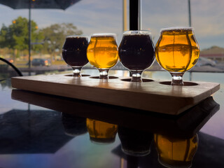 Small curved tulip shaped sample glasses of various ale beers with foam on the top. The samples are stout, red ale, pale ale, and IPA style beer on a wooden flight tray. The samples are on a table.
