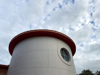 The exterior of a modern round building with a gold colored metal roof. The circular eave overhangs a grey colored panel wall. There's a small round window with glass and spacers. The sky is cloudy.