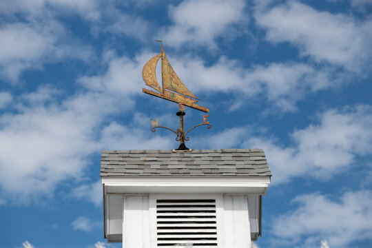 A rusty metal weather vane in the shape of a sailboat with the letters N, S, W, and E as directional markers below. The architectural ornament is on a cupola of a white wooden building. It has vents.