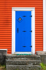 A bright blue wooden bifold traditional closed door with black wrought iron hinges, handle, and latch. The trim around the door is white. The vibrant red house has wooden clapboard siding and steps.  