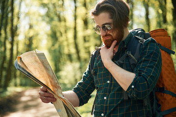 Old fashioned paper map in hands. Bearded man is in the forest at daytime