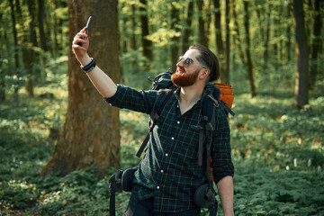 Phone in hand. Bearded man is in the forest at daytime