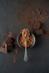 Spoon on a black bowl with cocoa and chocolate pieces on a dark concrete table. Top view with space for text.