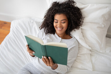 Hispanic woman is lying on a bed, engrossed in a book she is holding in her hands and smiling. The...