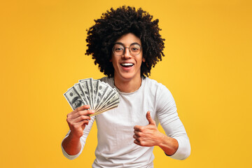Saving money concept. Happy african-american guy holding bunch of money and showing thumb up...